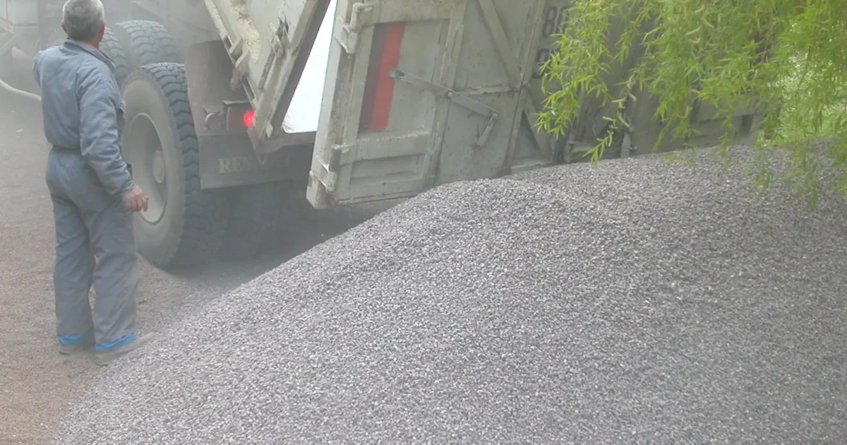 A dump truck tipping a load of gravel into a garden while a man watches