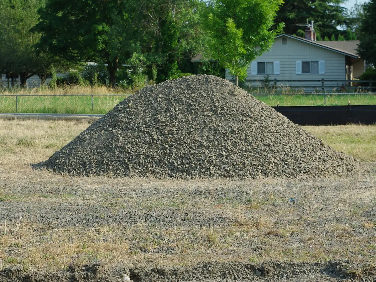 A large pile of gravel on bare ground with a house behind it