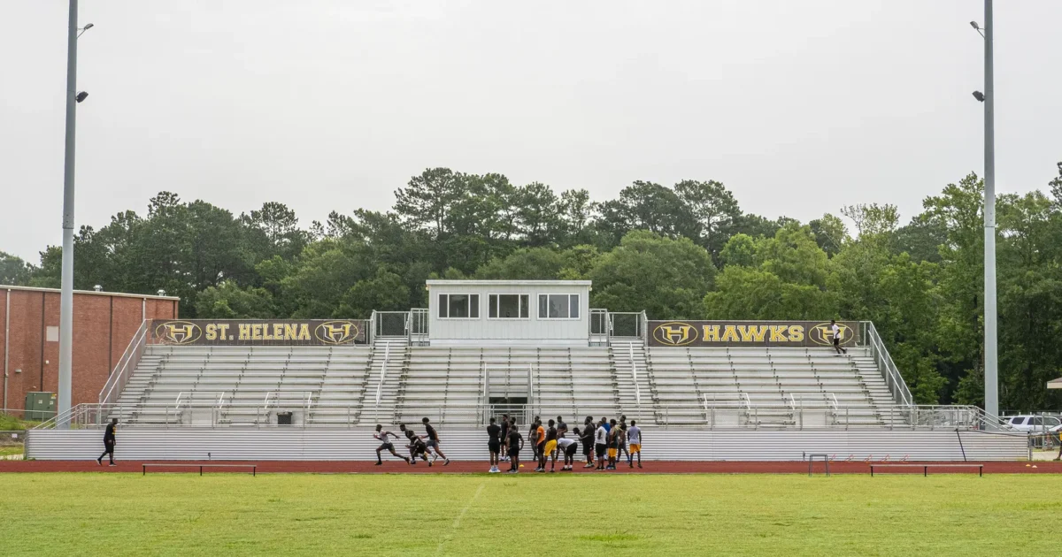 A high school football stadium with bleachers and a press box, with players on the field