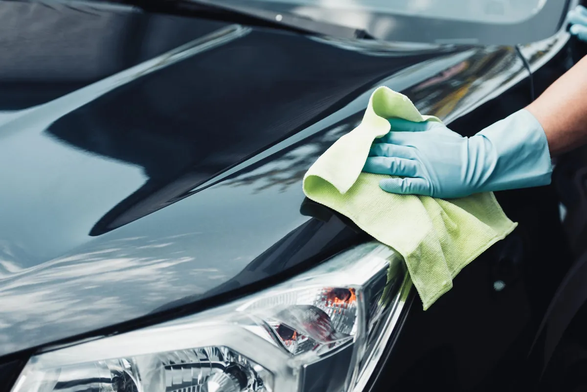 A gloved hand wiping a dark car with a green microfiber towel