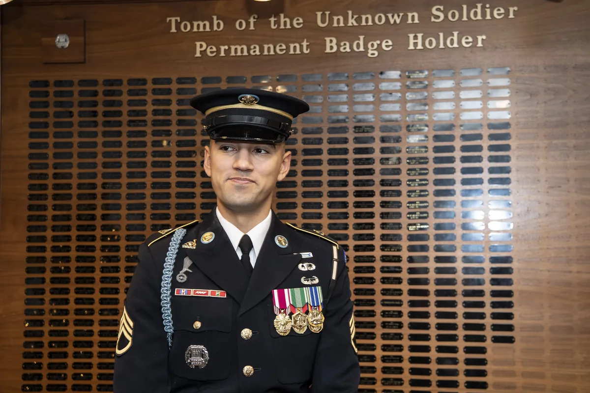 A staff sergeant in dress uniform in front of the board listing the permanent Tomb Guard badge holders