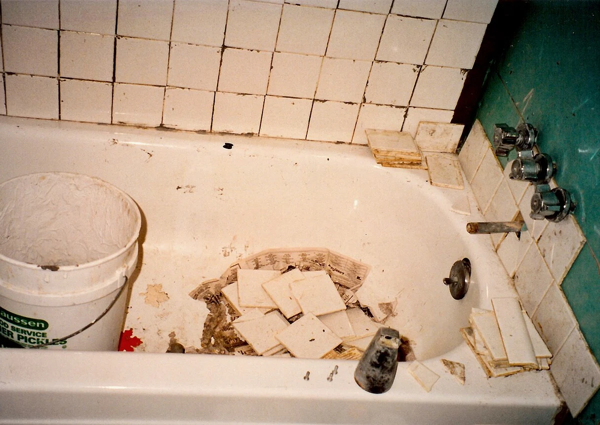 Tiles removed from the wall around a bathtub, with debris in the bath.