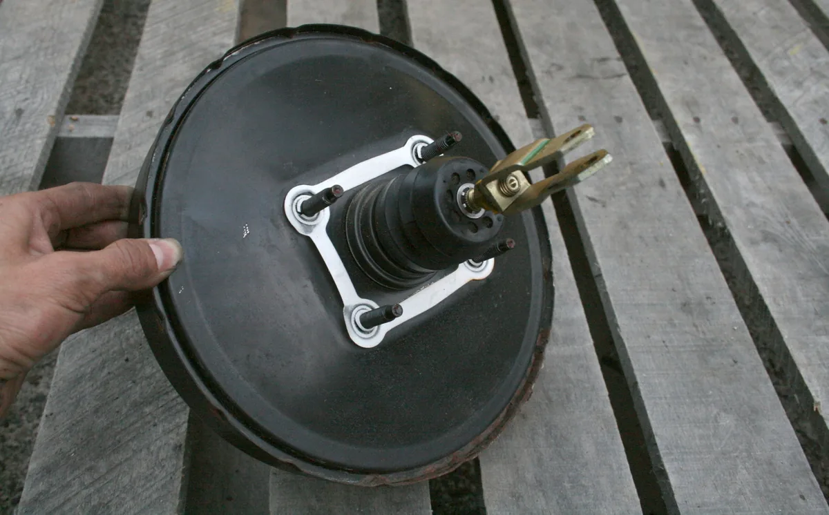 A round black brake booster held in a hand over a wooden bench, with its mounting studs and pushrod