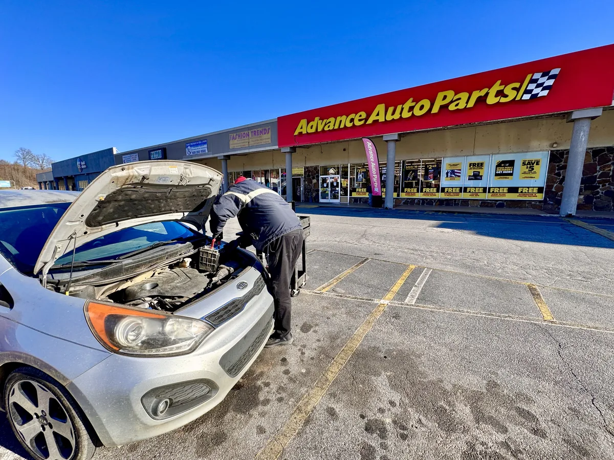 A parts store employee in a parking lot working on the battery of a car with its hood open