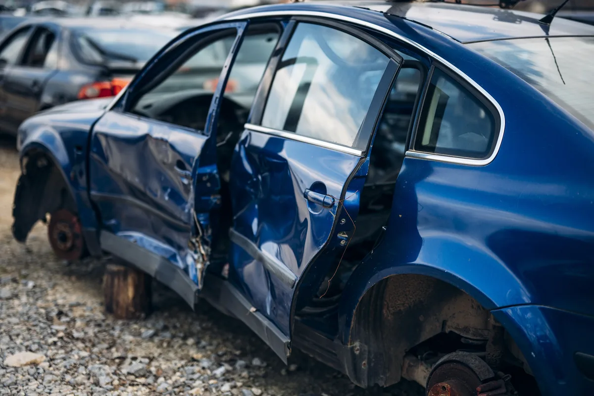 A blue car in a salvage yard with deeply dented side doors and missing wheels.
