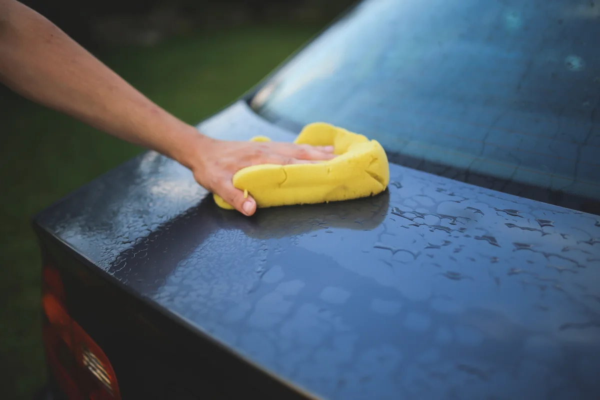 A hand using a yellow sponge on a wet car exterior.