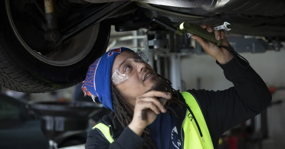 An automotive helper examines the underside of a raised vehicle with a flashlight.