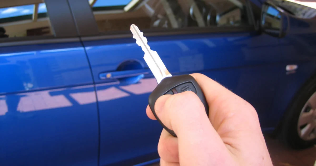 A hand holding a metal car key with remote buttons in front of a blue car.