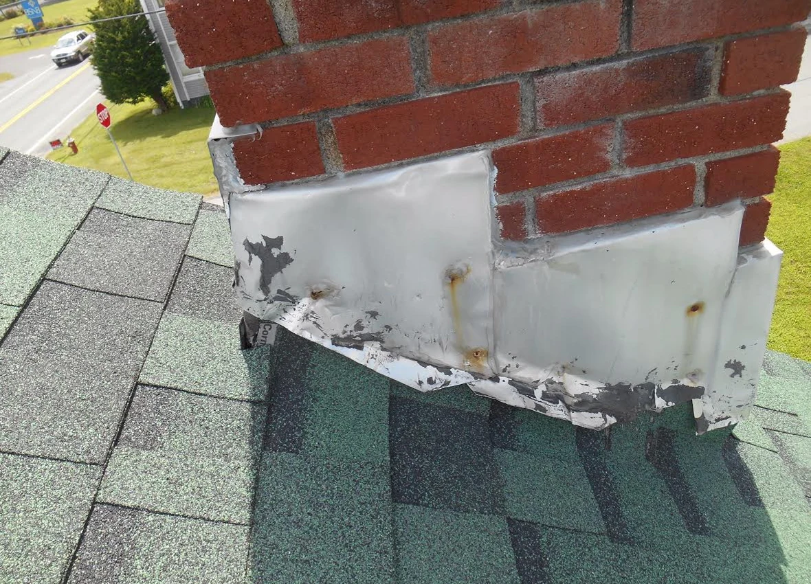 Old metal flashing around the base of a brick chimney on a shingle roof.