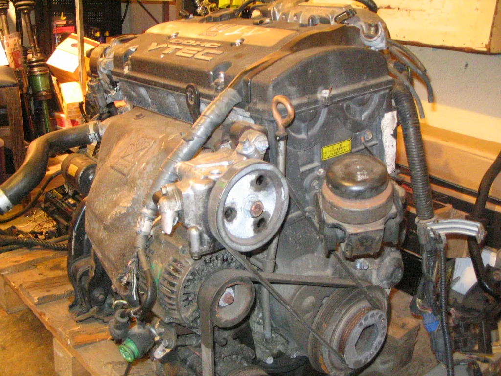 A Honda four-cylinder engine on a wooden pallet in a garage, seen from the belt side, with the crankshaft pulley at the bottom right