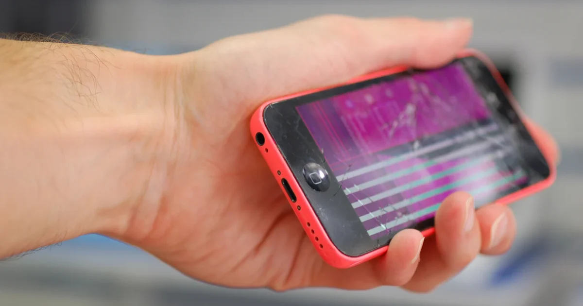 A hand holding a pink iPhone 5c with a damaged display, shown sideways.