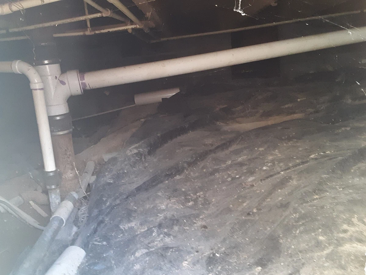 Crawl space seen through an access panel, with an old plastic vapor barrier partly covering the dirt floor and white pipes above.
