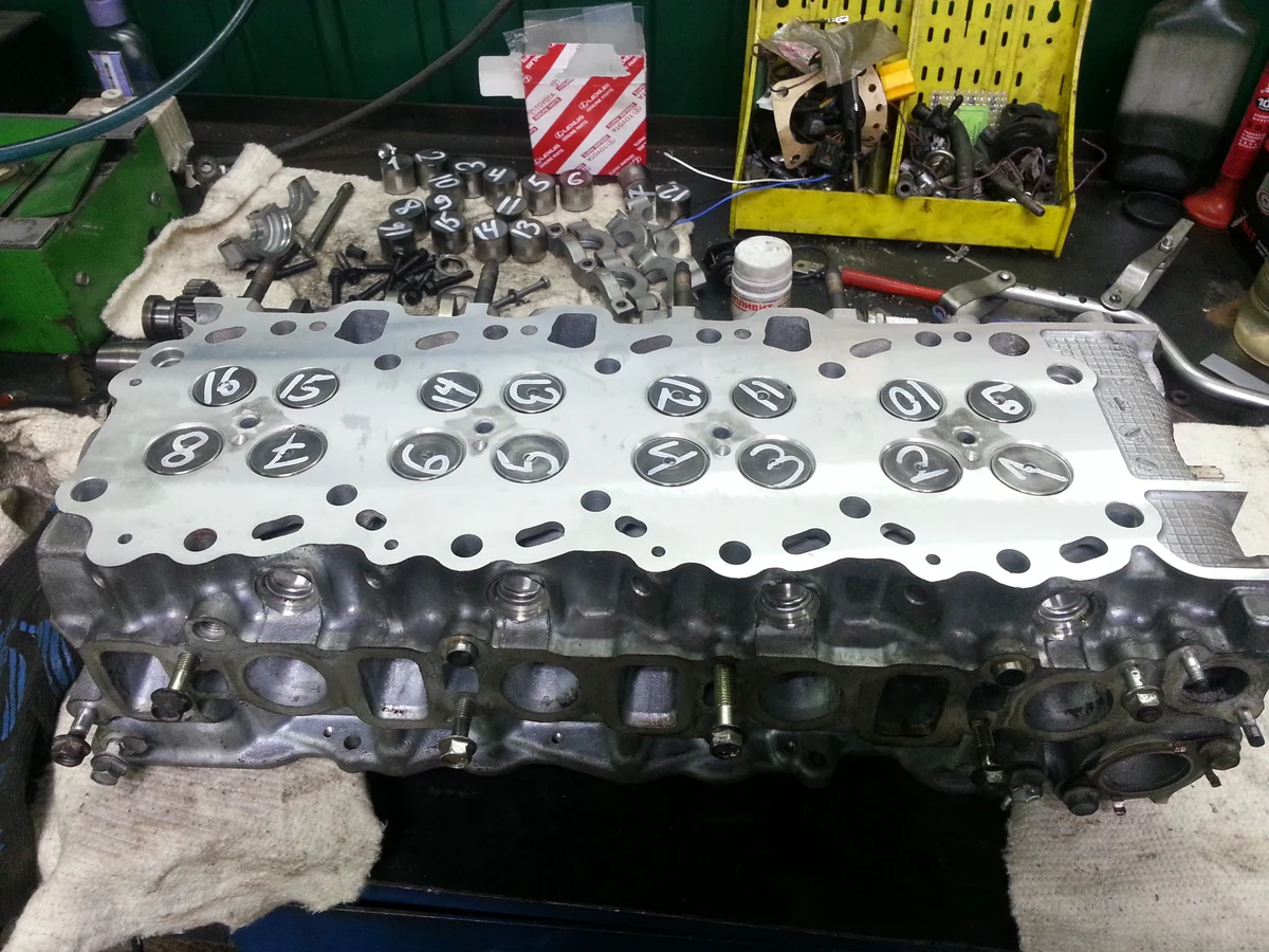 A removed Toyota 1KD-FTV cylinder head on a workbench, with numbered valves and dismantled parts behind it.