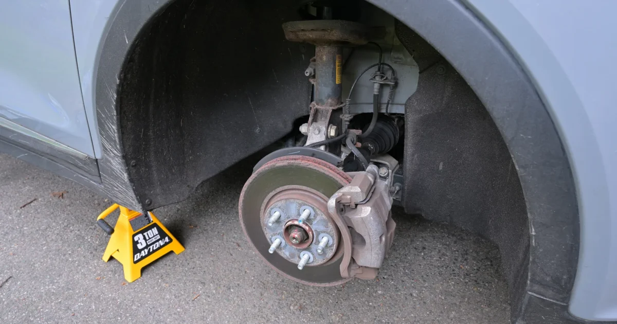 The front wheel well of a silver car with the wheel off on a jack stand, showing the brake rotor and the caliper clamped over it
