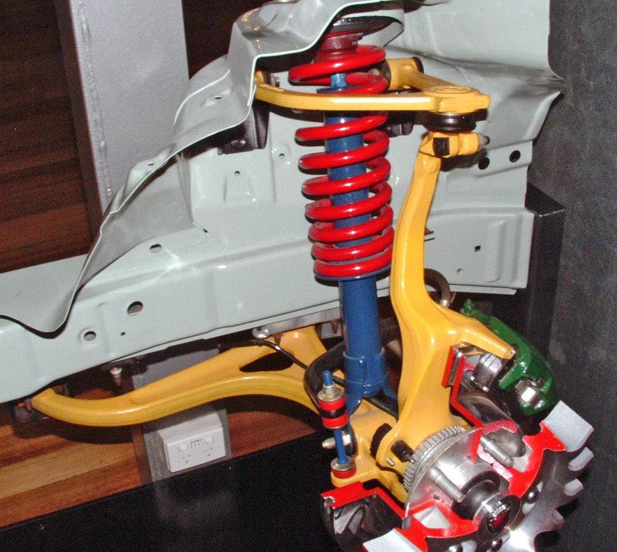 A cutaway display of a car front corner with yellow upper and lower arms, a red coil spring and the wheel hub