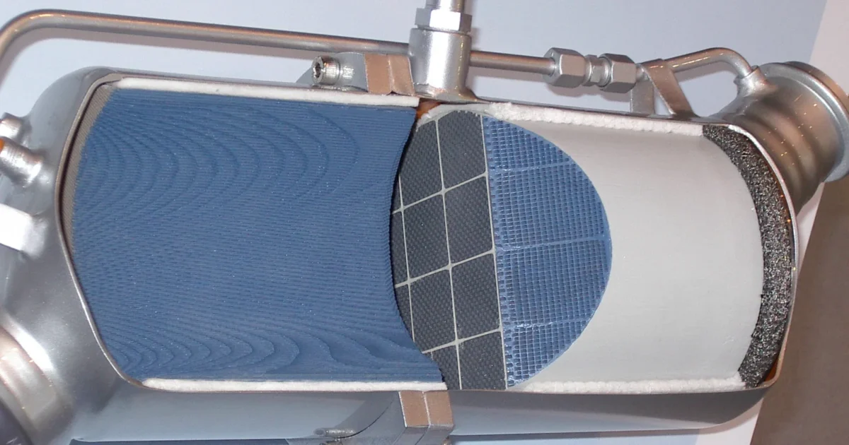 A steel exhaust canister cut open to show a blue oxidation catalyst and a grey honeycomb filter inside, with sensor pipes on top