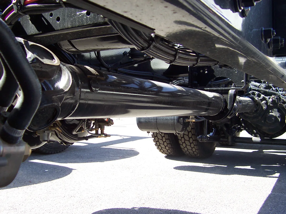 The long black drive shaft under a truck, seen from ground level between the wheels