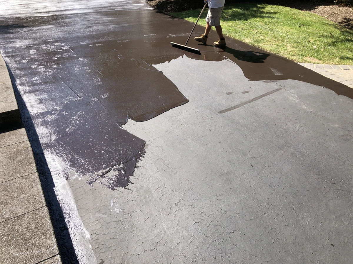 Worker applying a dark sealcoat over an existing asphalt driveway.