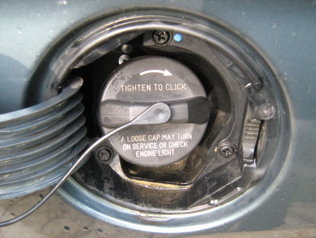 A gray gas cap in an open fuel door, printed with Tighten to click and A loose cap may turn on service or check engine light
