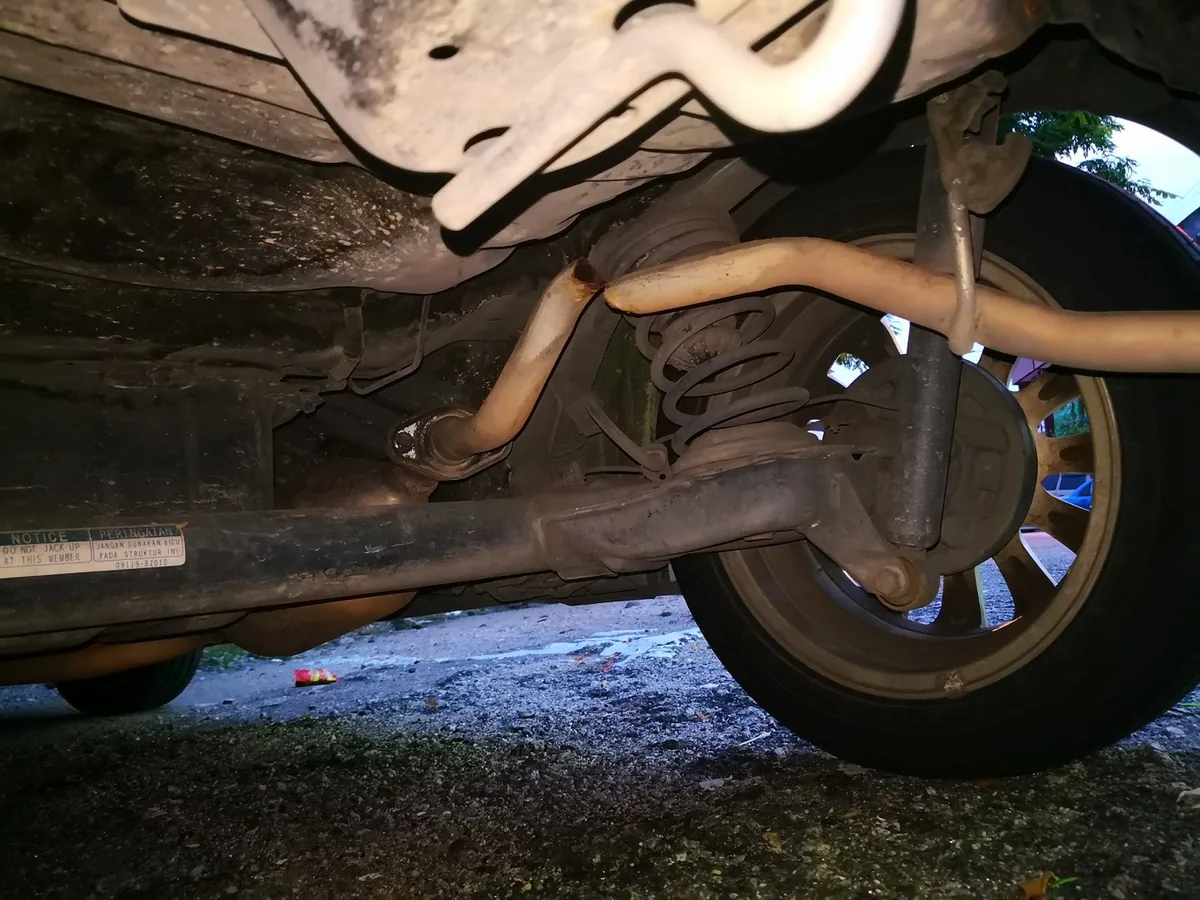 The rusty exhaust pipe under a small car, broken at a joint in front of the rear wheel
