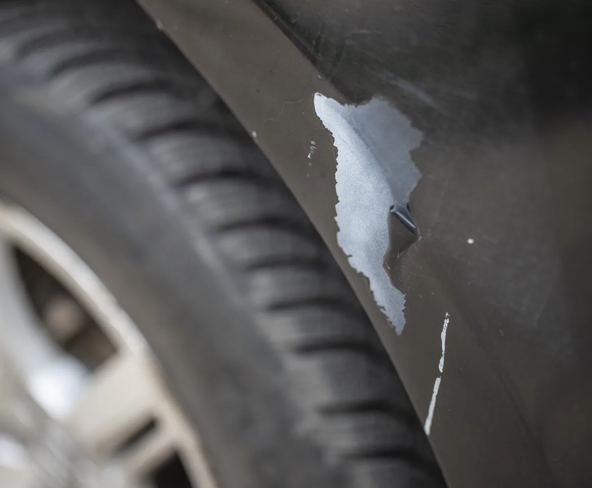 The black fender of a car above the front tire, with long scratches and a patch where the paint is missing