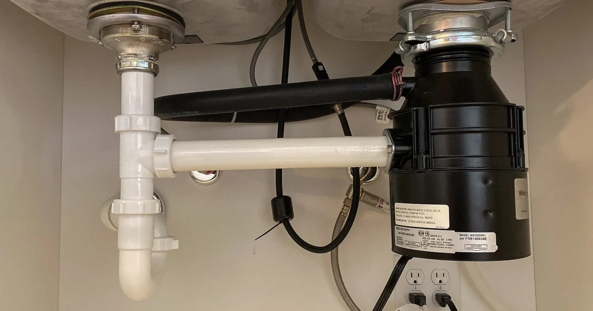 A black garbage disposal mounted under a double sink beside drain pipes.