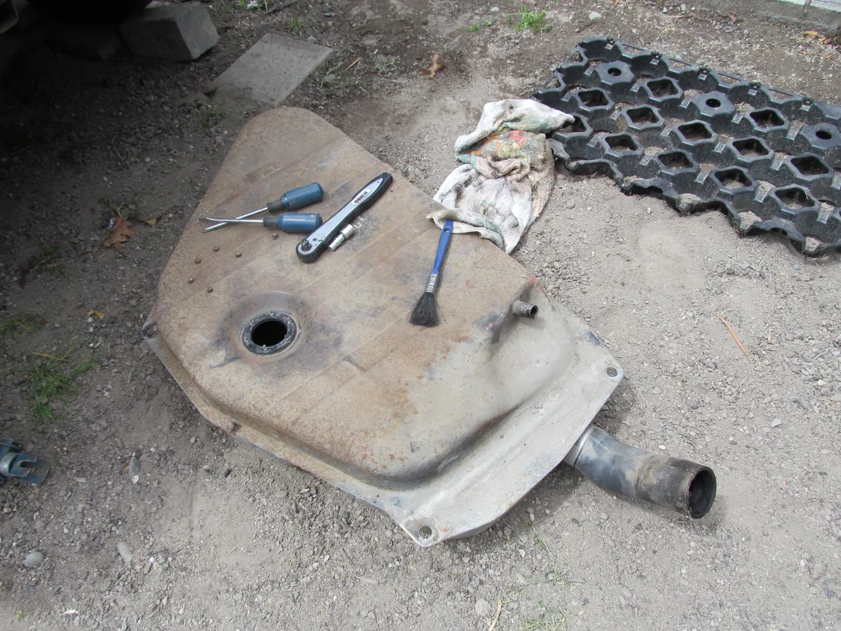 A dented metal car fuel tank lying on dirt with its filler pipe stub, next to screwdrivers and a rag
