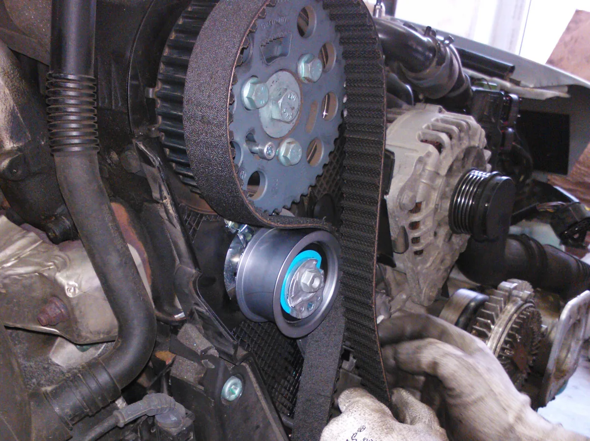Gloved hands working on a black toothed timing belt running over the pulleys at the front of an engine