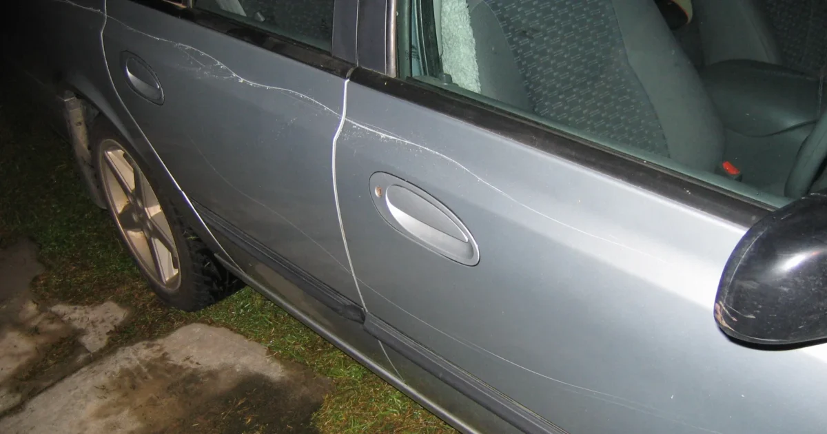 The side of a silver car with long thin scratches across both doors, where someone dragged a key along the paint