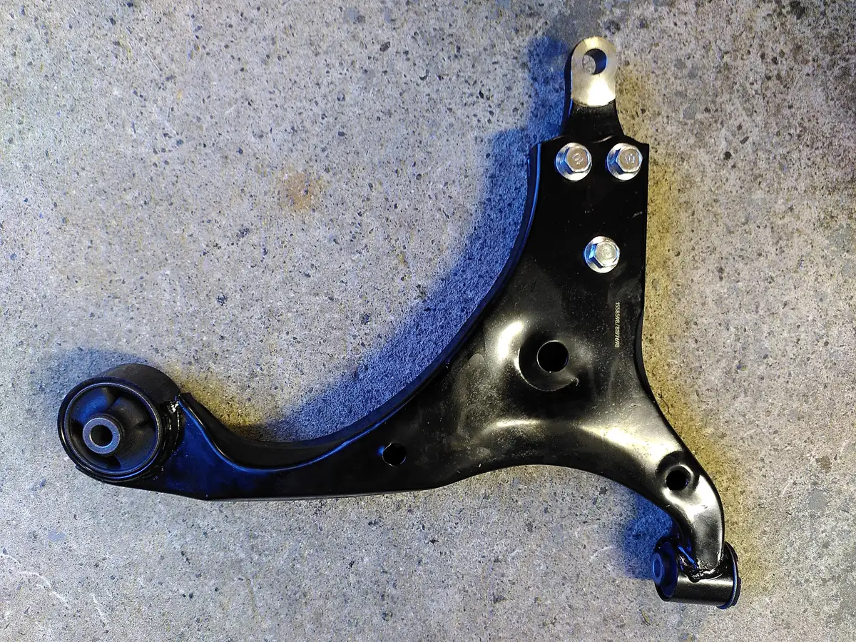A new black lower control arm lying on concrete, with round rubber bushings at two of its ends