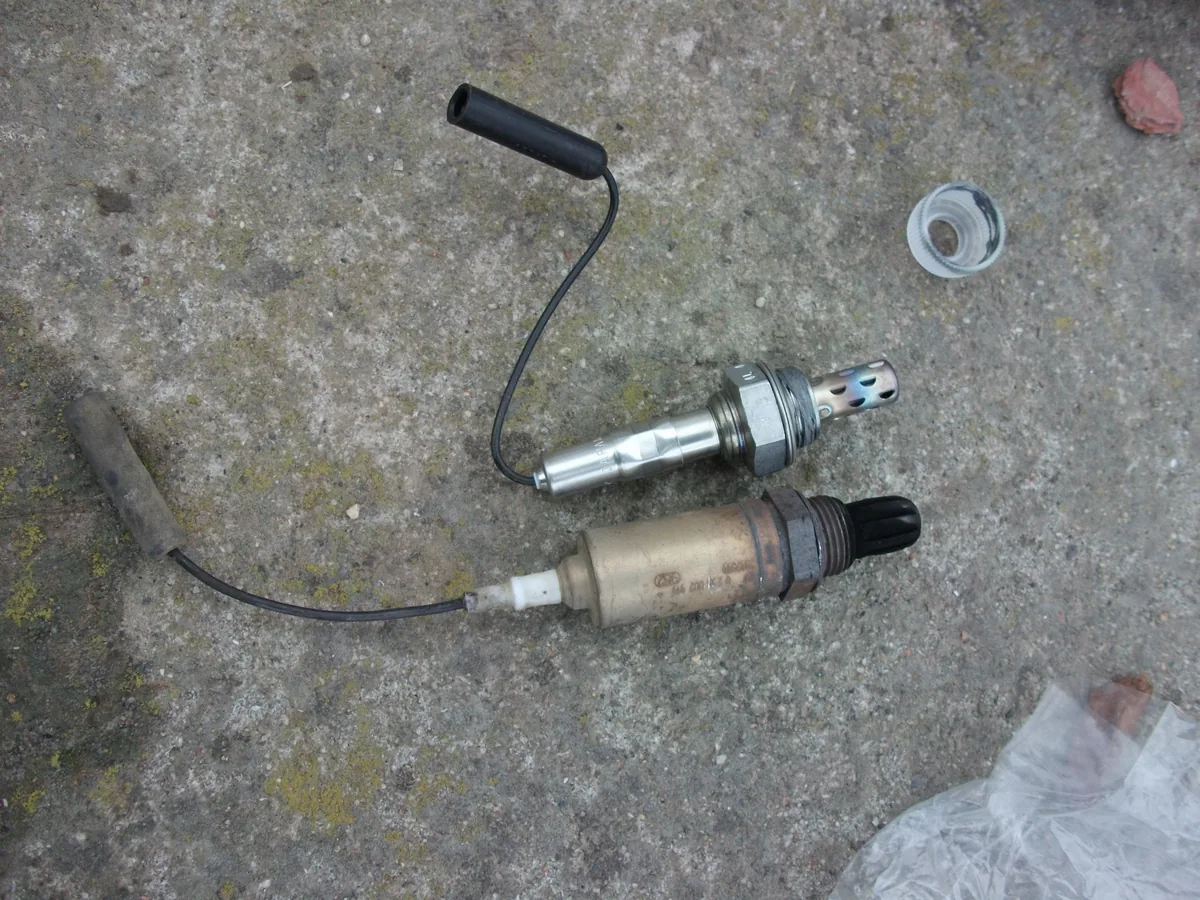 Two oxygen sensors on a garage floor: a shiny new one with a black wire and plug at the top, and the old brown one with a gray wire below it
