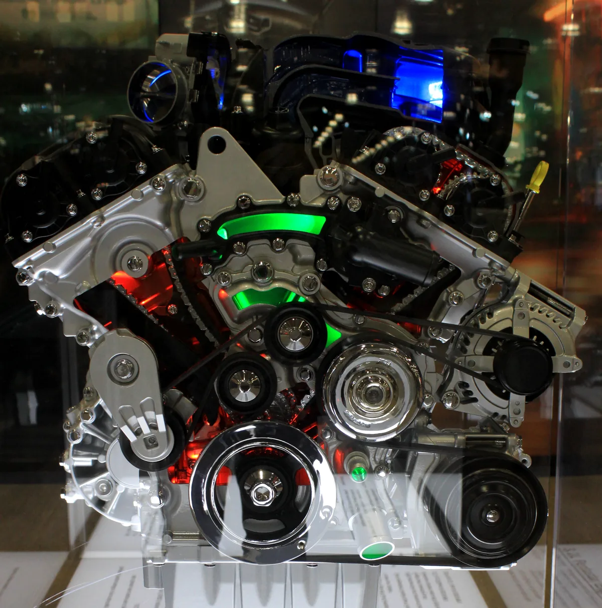A Pentastar V6 display engine with parts of its front cut open and lit, showing the timing chains and their guides