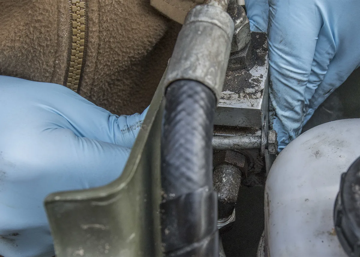 Gloved hands holding a black rubber hose with a metal fitting next to a white fluid reservoir