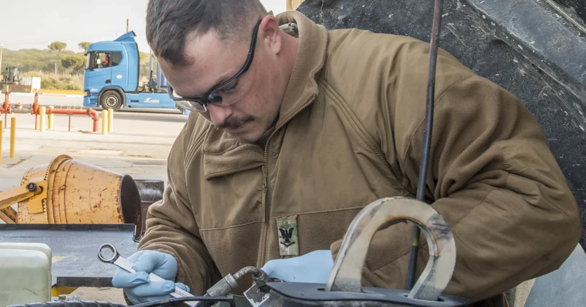A mechanic in glasses and gloves working with a wrench under the open hood of a military vehicle