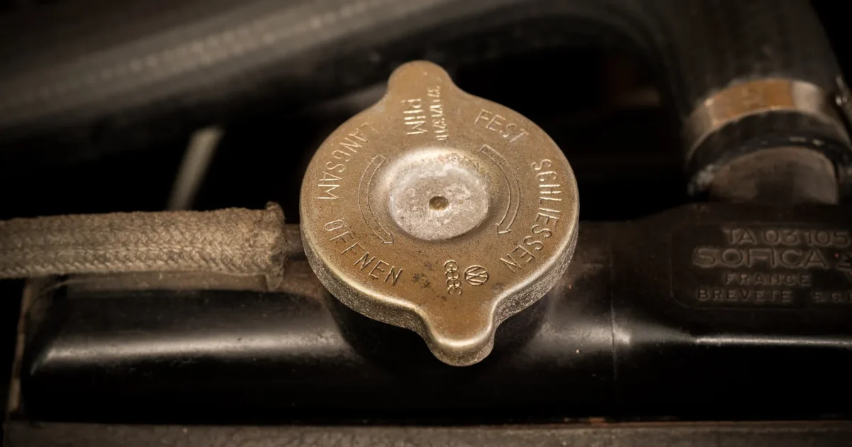 A round metal radiator cap on the filler neck of a car radiator, with its warning text stamped around the top and the radiator hose behind it