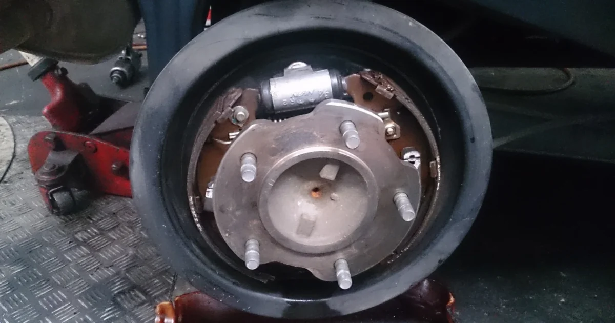 The rear wheel of a car raised on a jack with the wheel off, showing the round brake drum with the wheel studs and the brake parts behind it
