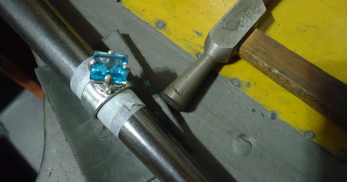 A blue topaz ring on a tapered jewelry mandrel beside a hammer.
