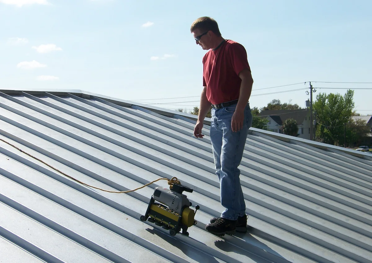 A worker beside a roof-seaming machine on a metal roof.