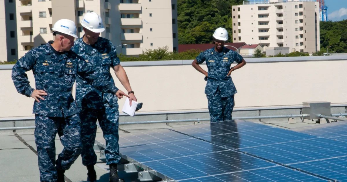 U.S. Navy officials in hard hats inspecting rows of blue solar panels on a school roof in Japan.