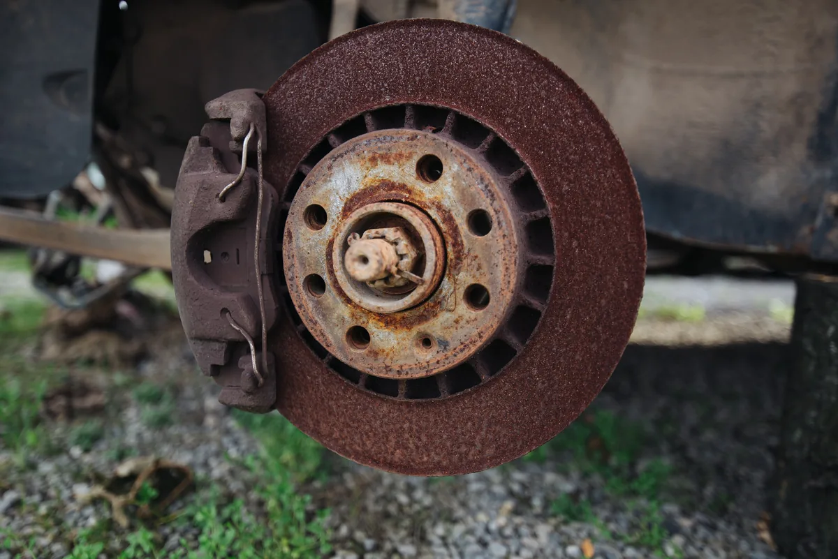 A brake rotor covered in rust with its caliper, on a car parked on gravel with the wheel off