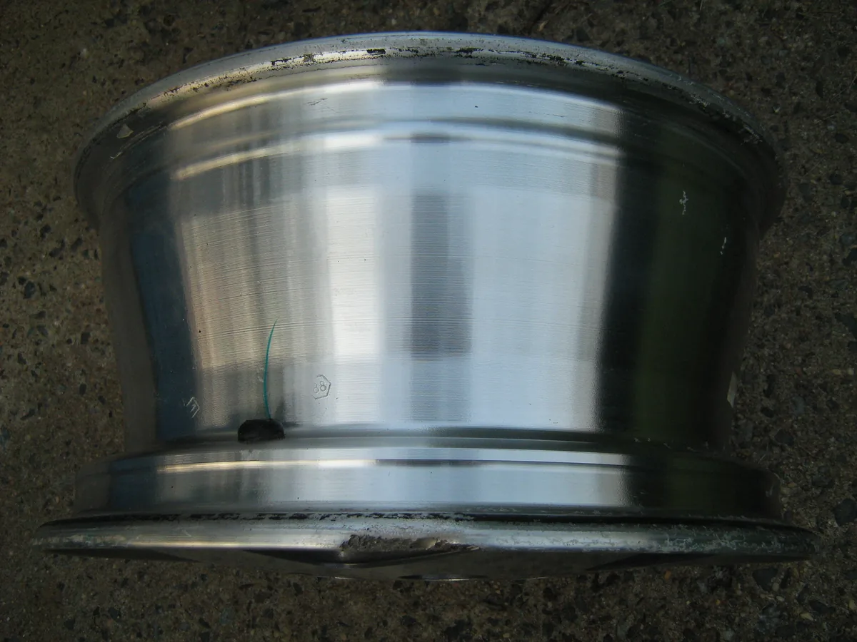 A bare silver alloy wheel seen from the side on a gravel floor, with scratches along the lower rim lip