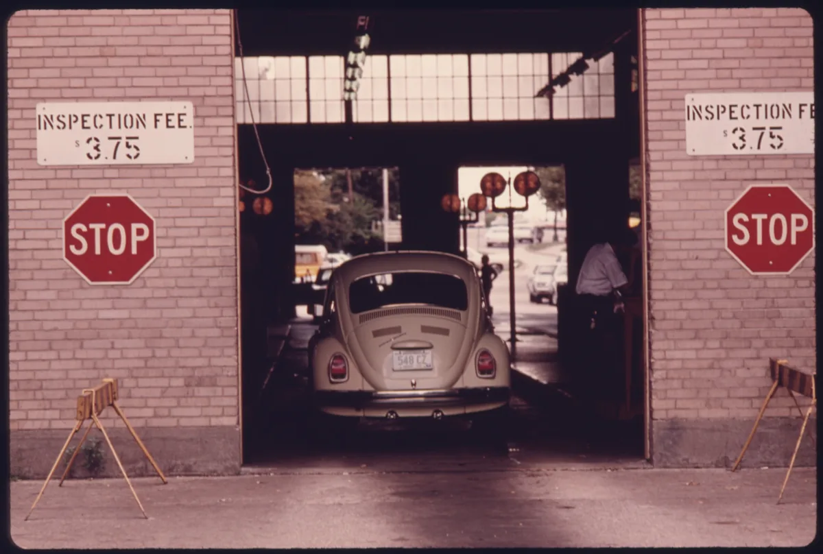 A Volkswagen Beetle entering an inspection lane between two stop signs, with signs on the wall that show an inspection fee of $3.75