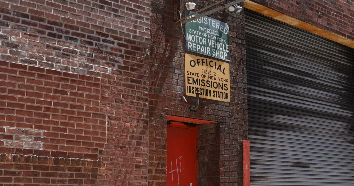 A brick repair shop with a red door and a roller shutter, under signs for a registered motor vehicle repair shop and an official New York State emissions inspection station