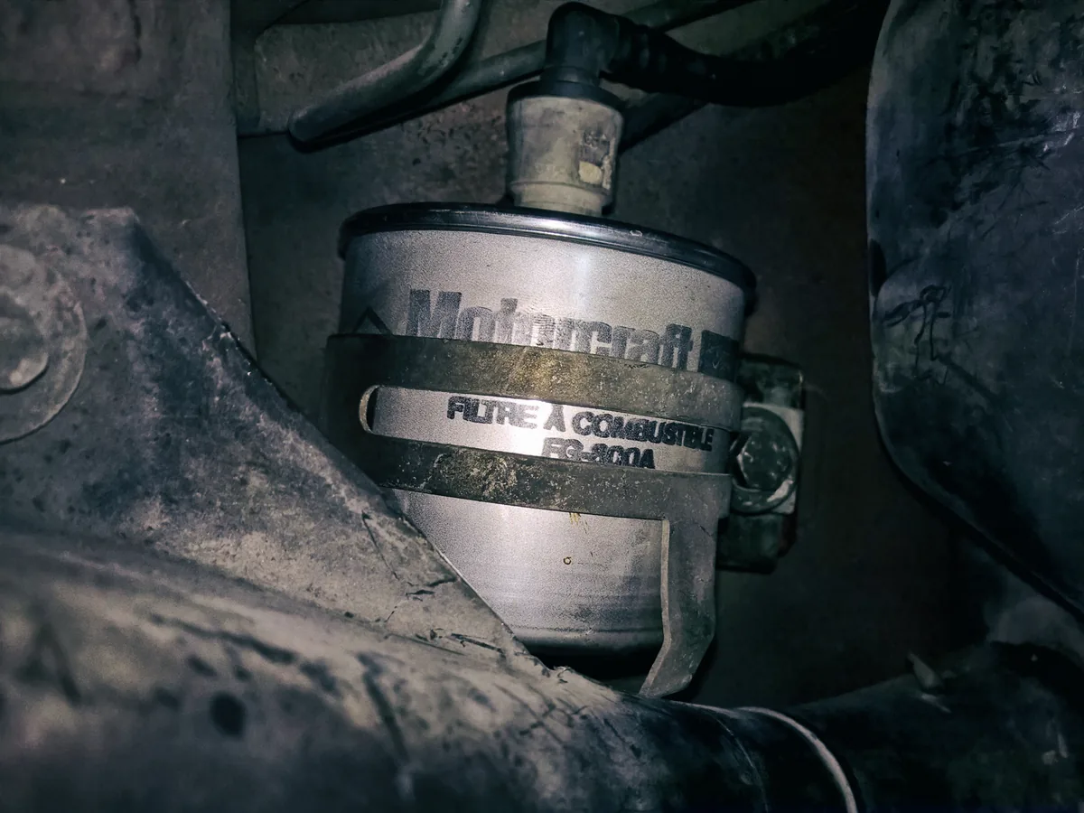 A metal fuel filter clamped under a Ford Escort with steel fuel lines running into both ends