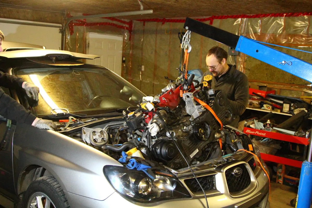A man in a home garage lifting a Subaru engine out of a silver Impreza with a blue engine crane