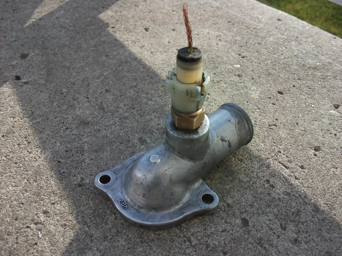A metal thermostat housing off a Toyota Corolla on a concrete step, with a temperature switch and its wire screwed into the top
