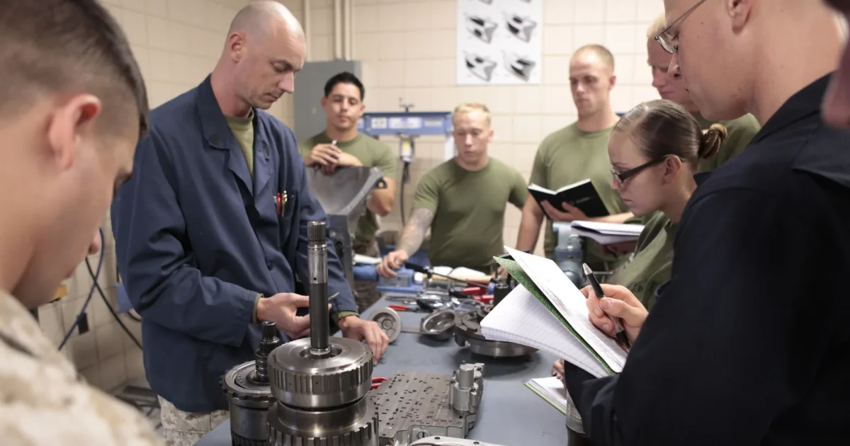 An instructor in blue overalls with students at a workbench covered with the parts of an automatic transmission