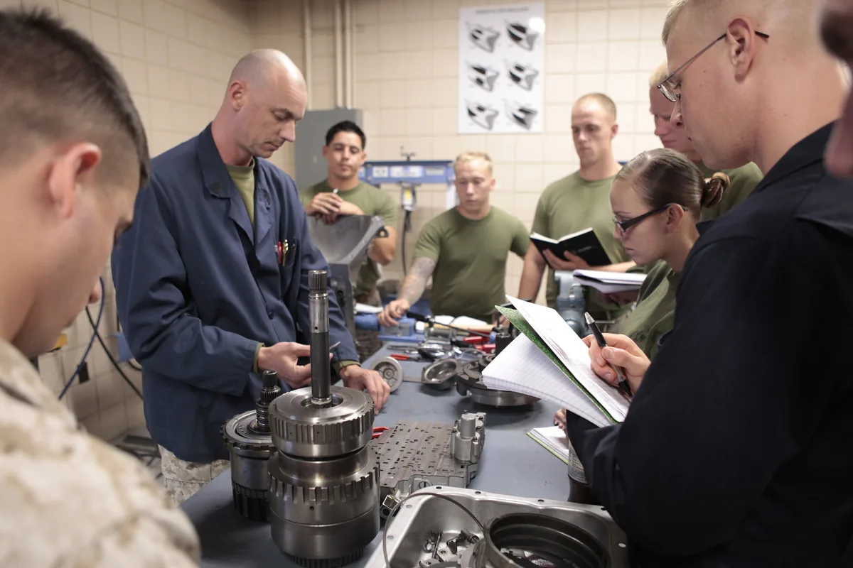 A Marine Corps instructor in blue coveralls with students around a workbench where an automatic transmission lies taken apart