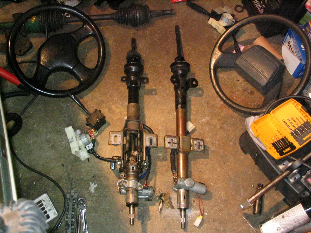 Two steering columns from Nissan cars laid on a garage floor, with steering wheels and their long shafts beside them