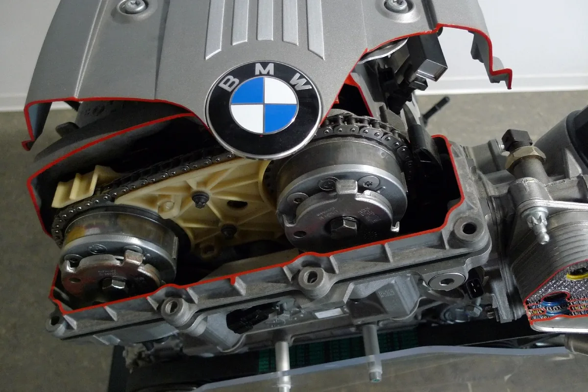 A cut open BMW six cylinder engine showing two round cam phasers on the ends of the camshafts and the timing chain around them